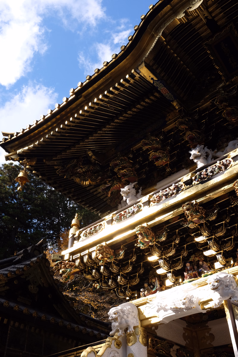 A close-up view of an ornate, dark temple roof covered in intricate gold decorations against a bright blue sky with white clouds.