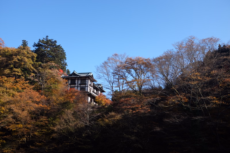 A traditional Japanese temple building is nestled among colorful autumn trees under a clear blue sky.