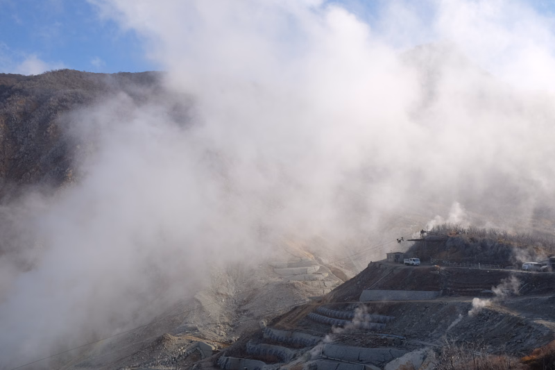 Thick white steam rises from the slopes of a volcanic mountain, obscuring parts of the rugged landscape.