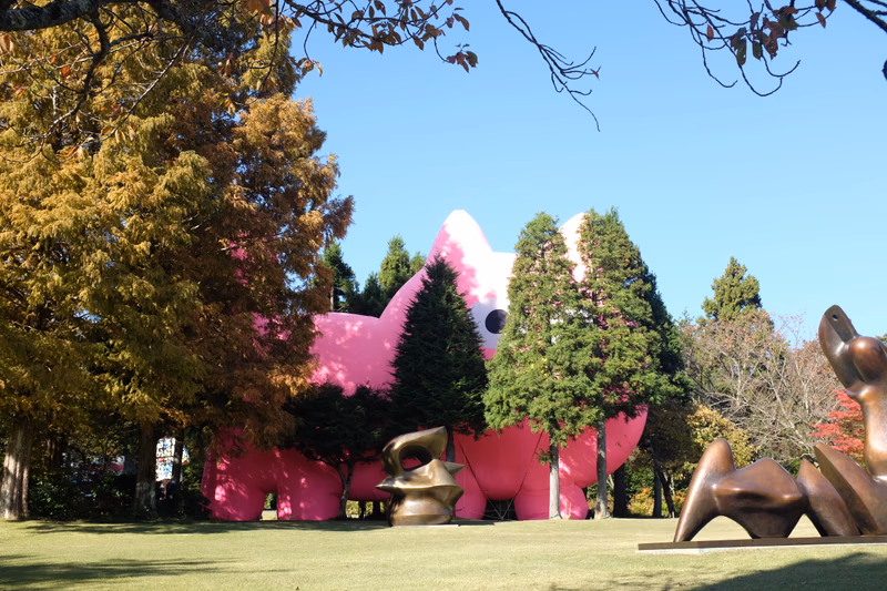 A photograph of a pink elephant sculpture in a park with trees and a bronze sculpture in the foreground.