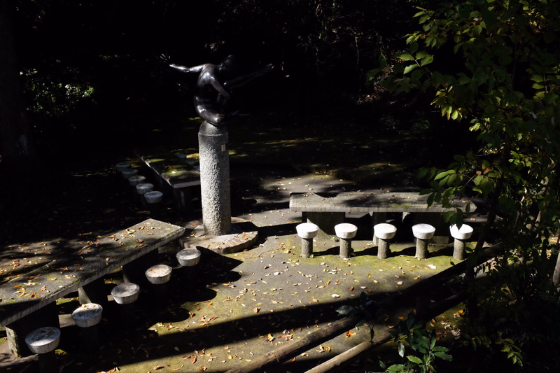 A dark bronze sculpture of a figure stands on a pedestal in a shaded garden area with stone steps.