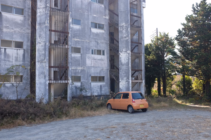 An orange car is parked on a gravel lot in front of a tall, weathered concrete apartment building surrounded by trees.