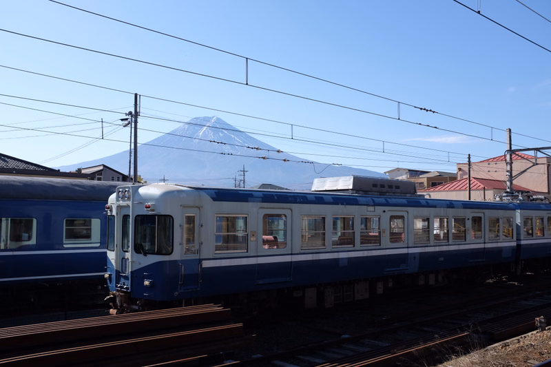 A blue and white train travels on tracks with Mount Fuji visible in the background under a clear blue sky.
