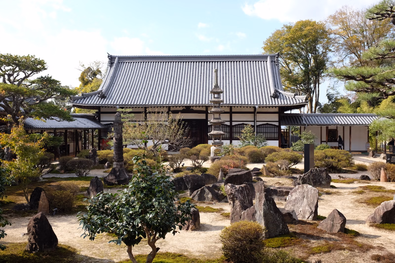 A traditional Japanese temple building with a dark tiled roof is surrounded by a meticulously maintained garden featuring rocks and trees.