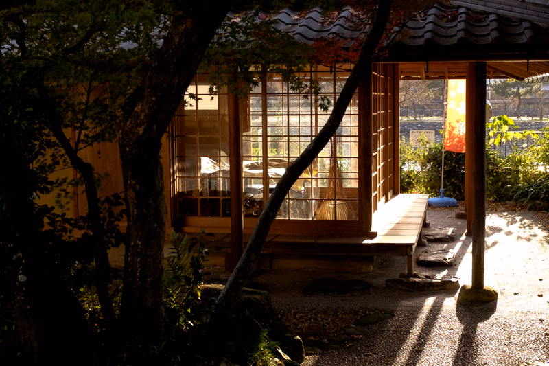 A traditional wooden tea house with glass windows sits in a shaded garden area, featuring a covered porch.