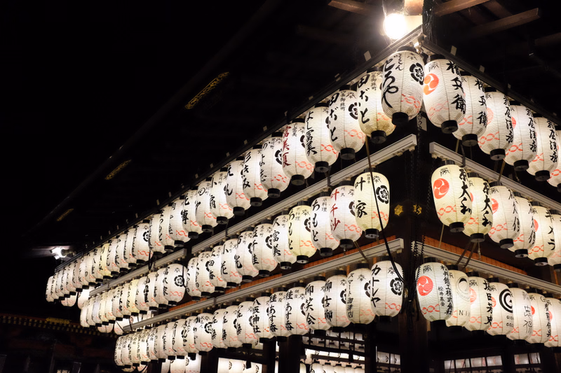 A display of numerous white paper lanterns hanging under a dark structure, some featuring Japanese characters.