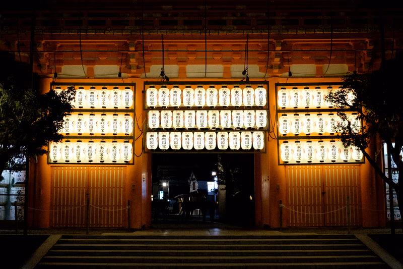 A nighttime view of a traditional Japanese gate illuminated with lanterns, with people walking through the entrance.