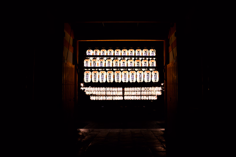 A row of illuminated paper lanterns hangs in a dark entryway, possibly leading to a Japanese festival or shrine.