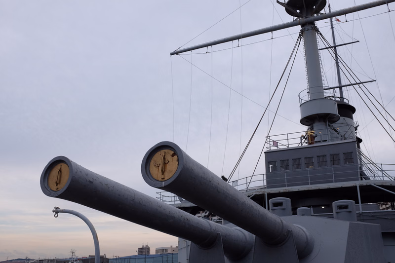 Two large naval guns are prominently featured on the deck of a warship against a cloudy sky.