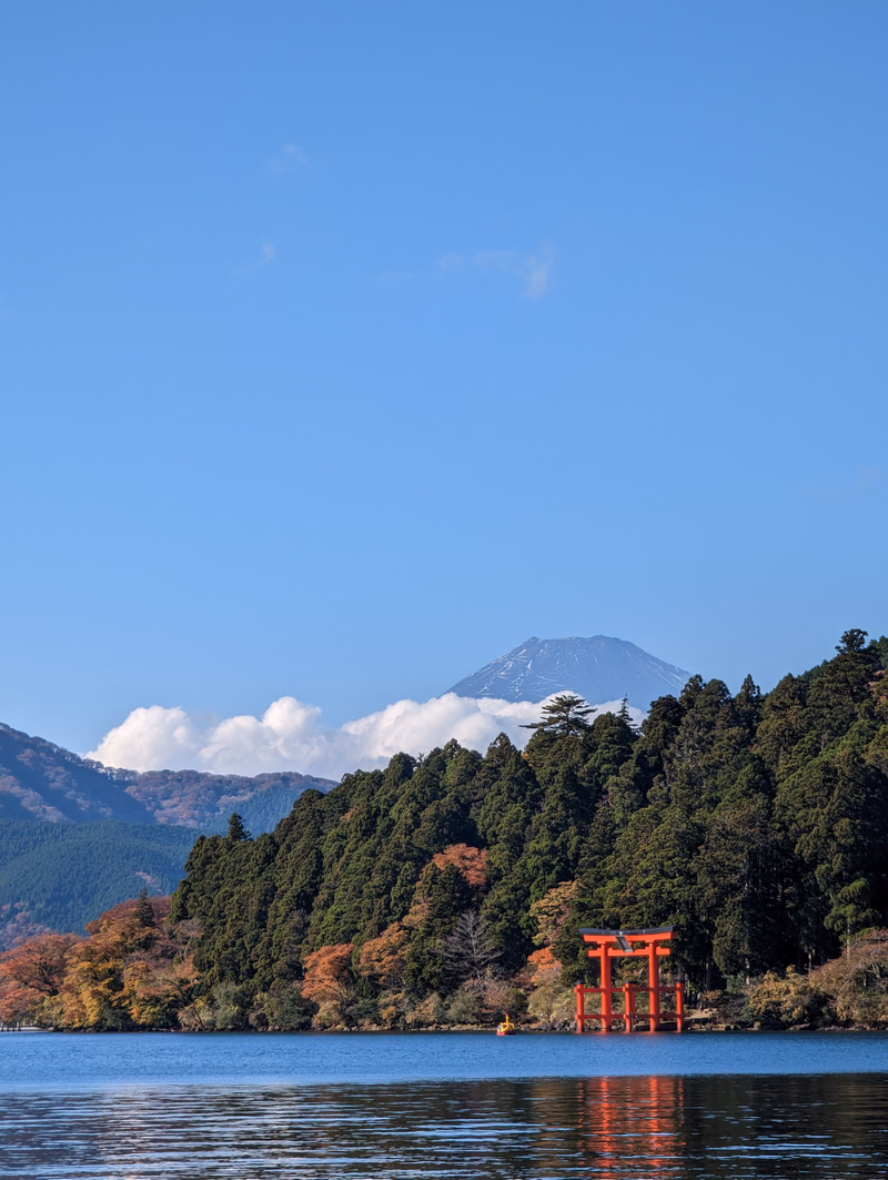 A red torii gate stands near the shore of a lake, with a snow-capped mountain visible in the background under a clear blue sky.