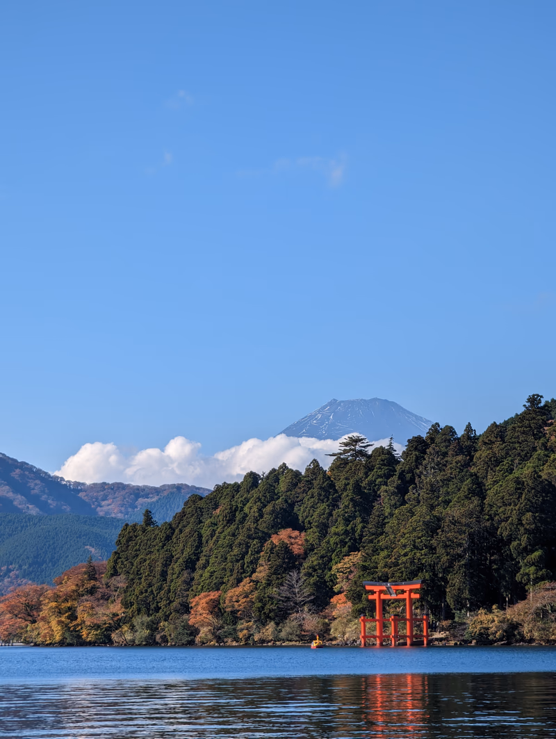 A serene lake with a red torii gate standing in the middle, surrounded by a dense forest, with a majestic mountain in the background under a clear blue sky.