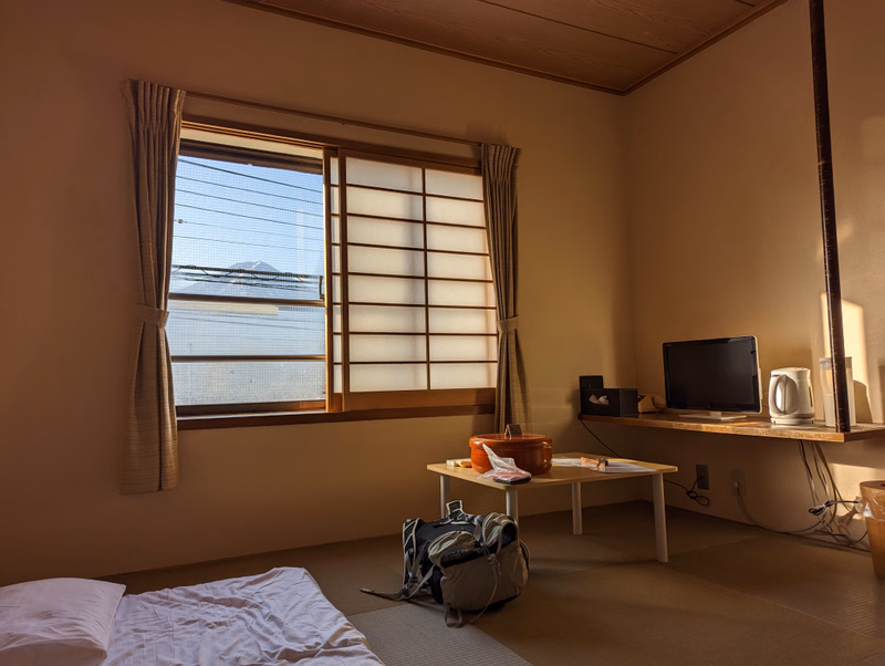 A bright, minimalist bedroom features a large window with blinds, a bed in the foreground, and a small wooden shelf with a television.