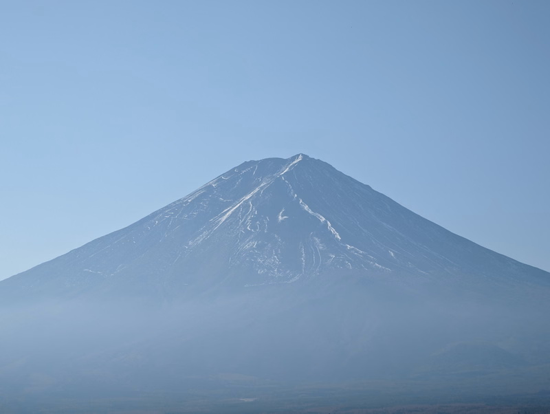 A large, snow-capped volcano rises against a clear, pale blue sky, partially obscured by low-lying fog.
