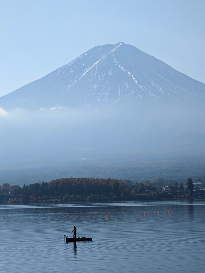 A person in a kayak paddles on a calm lake with a large, snow-capped mountain partially obscured by mist in the background.