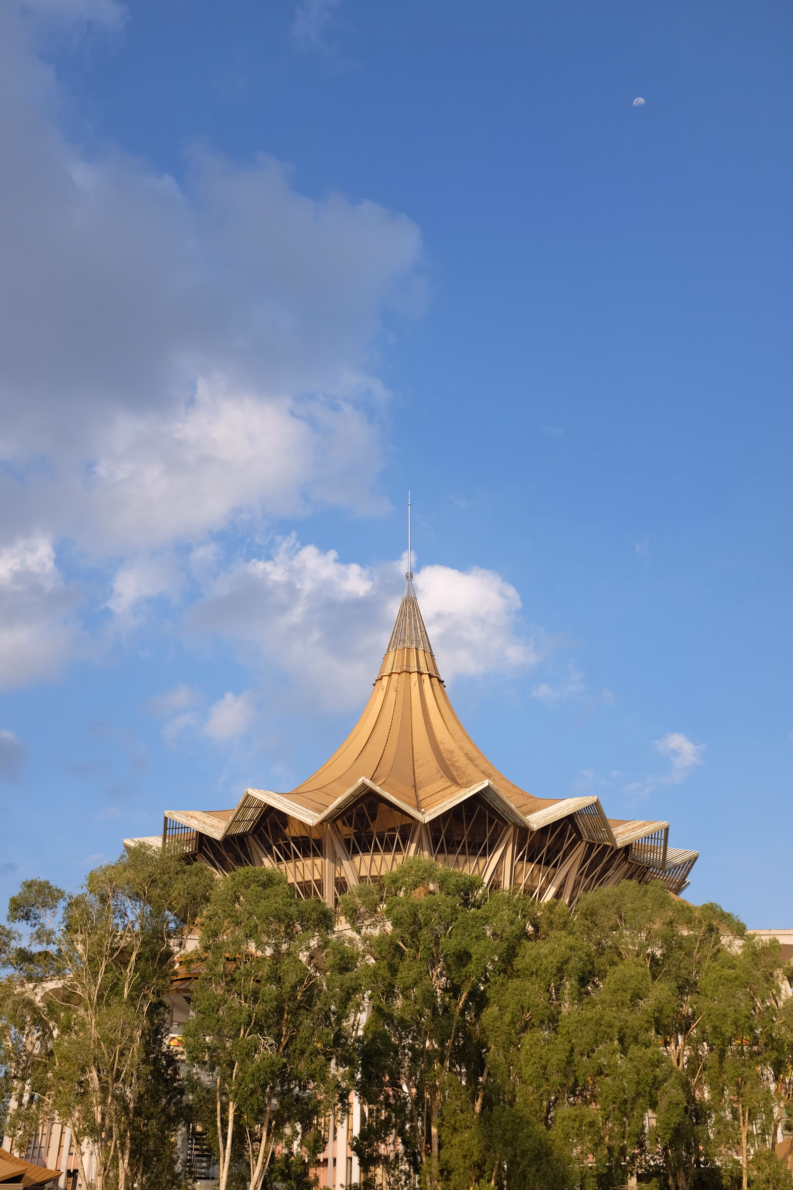 A distinctive cone-roofed building is partially visible above a dense canopy of green trees under a bright blue sky with scattered clouds.