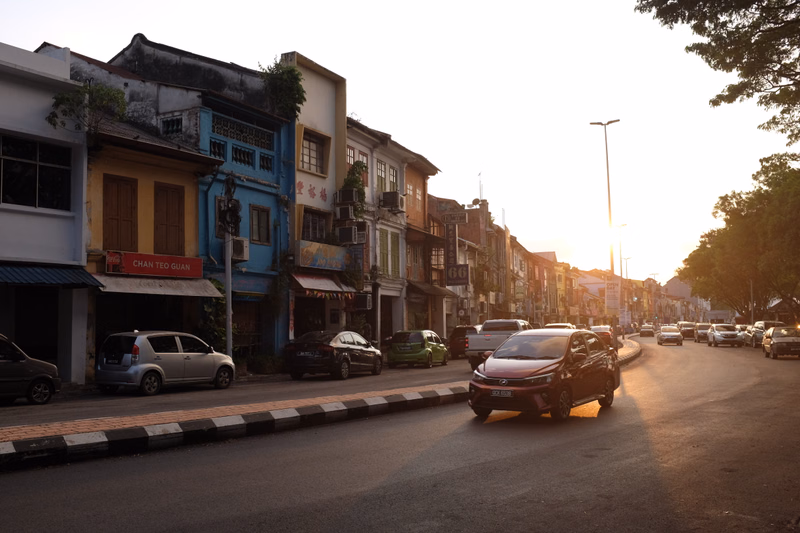 A row of colorful shophouses lines a street as cars drive past during sunset, with one sign reading "CHAN TEG GUAN".