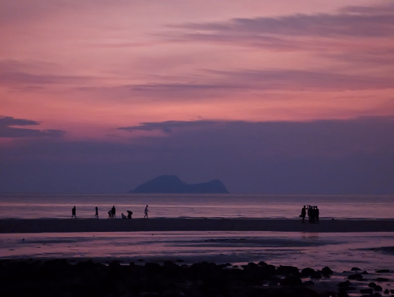 Silhouettes of people walking along a beach at sunset with a mountain visible on the horizon under a pink and purple sky.