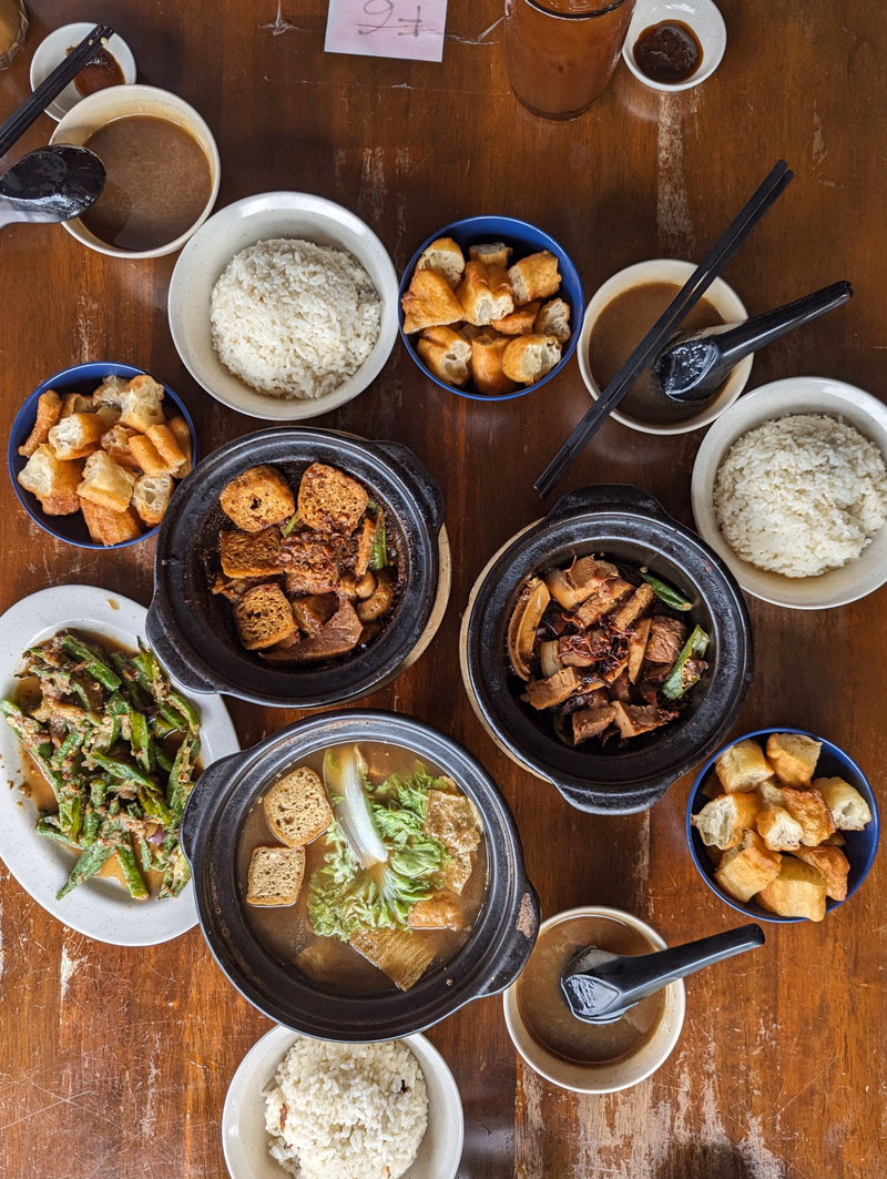 A variety of dishes, including rice, meat, buns, and soup, are arranged on a wooden table.