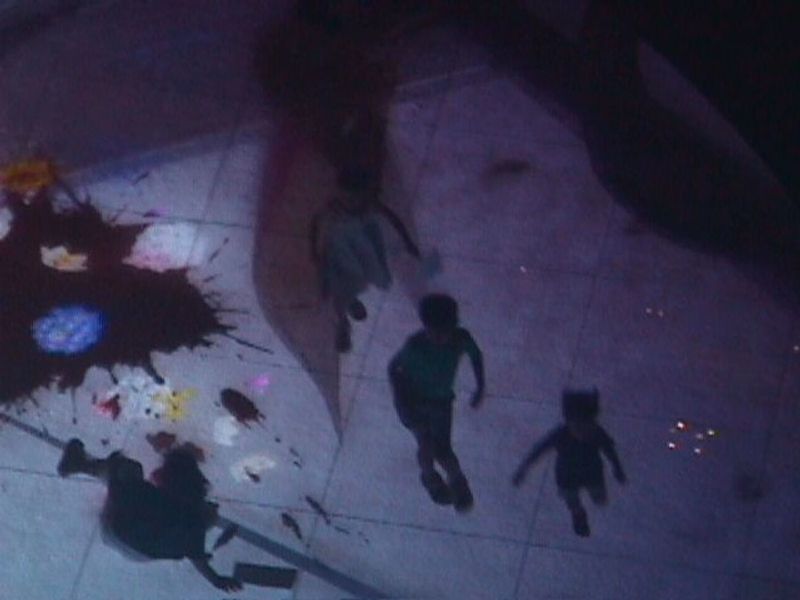 Several small figures are visible on a light-colored floor, with large, dark shadows cast across the scene.