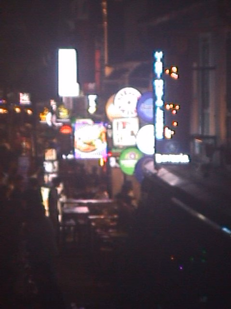 A dark city street at night is illuminated by various bright neon signs, including one that appears to read "Bavaria".
