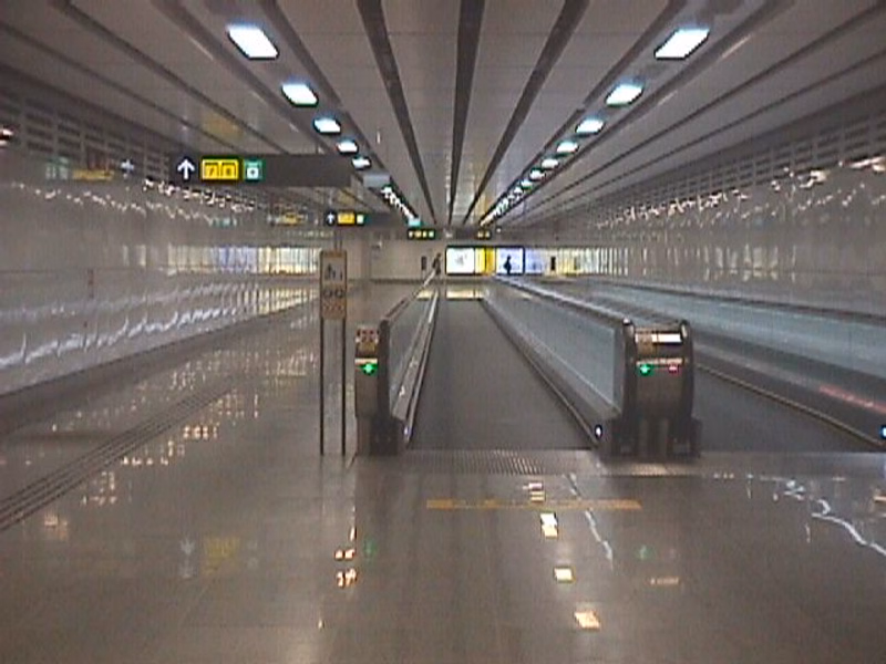 A modern, brightly lit train station platform with a train stopped at the end, featuring overhead lighting and digital signage.