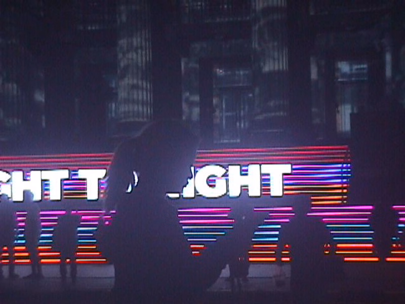 Silhouetted people stand in front of a large illuminated sign that reads "IGHT" against a dark city backdrop.