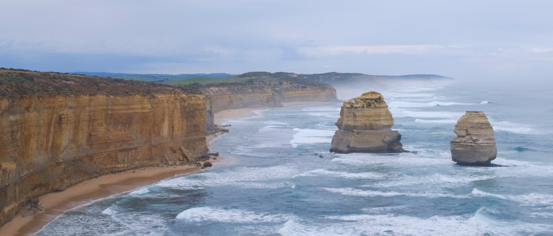 Dramatic sea stacks rise from the ocean near a rugged coastline with high cliffs and a sandy beach under an overcast sky.