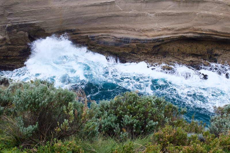 Large white waves crash against a layered, grey rocky coastline, with green coastal vegetation in the foreground.