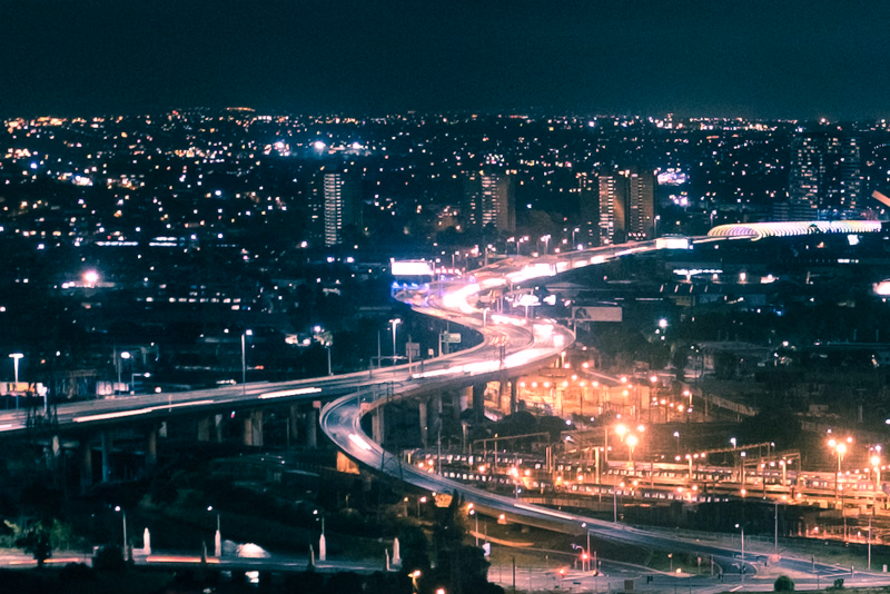 A wide nighttime view of a sprawling city illuminated by countless lights, featuring a curving highway interchange.