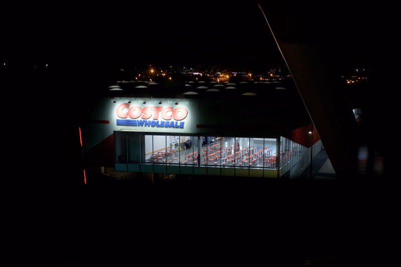 A large Costco Wholesale store is visible at night, with interior lights illuminating the warehouse.