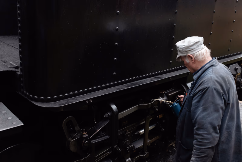 A man in a white hat is inspecting the mechanical parts of a black metal train.