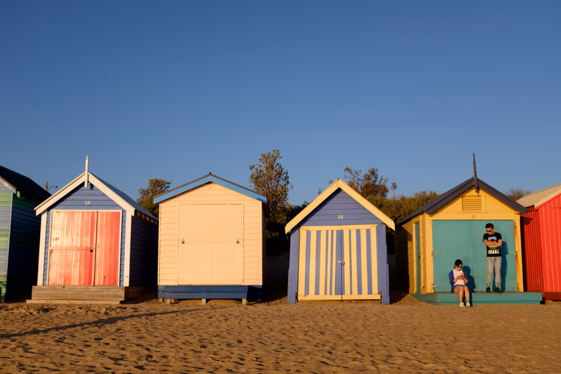 A row of colorful beach huts stands on the sand under a clear blue sky.