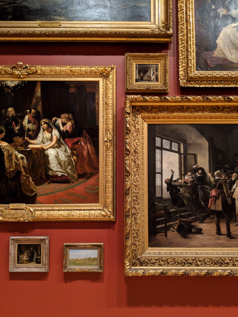 A collection of ornate, gilded picture frames displaying various paintings are mounted on a deep red wall in an art gallery.