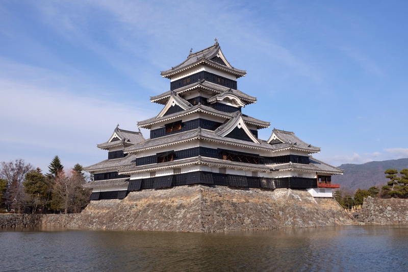 A traditional Japanese castle stands on a stone foundation surrounded by a moat under a bright blue sky.