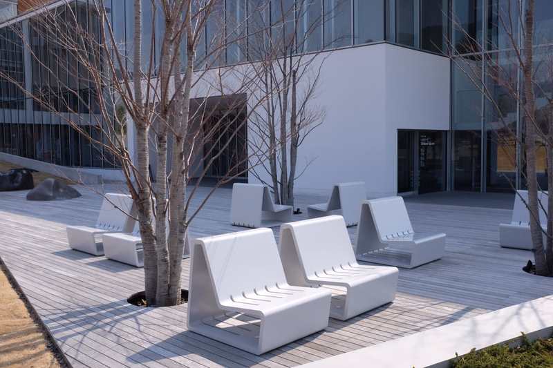 Several white modern lounge chairs are arranged on a wooden deck outside a contemporary glass building with bare trees nearby.