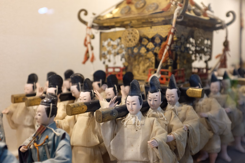 A group of traditional Japanese figures or statues are lined up in front of a large, ornate, golden shrine or float.