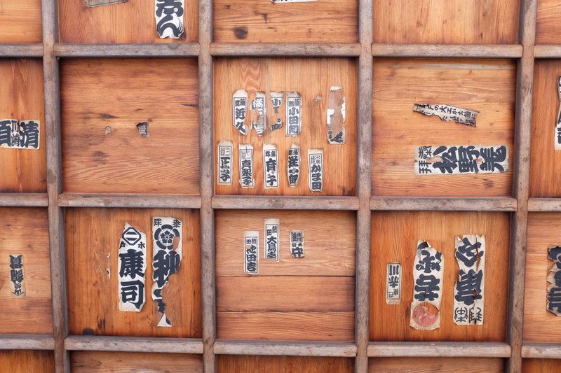 A close-up of a wooden panel covered with various handwritten and printed Japanese text, including characters like "魚" and "麺".