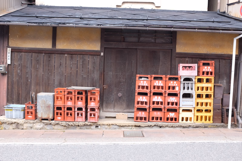 A row of colorful plastic storage crates is lined up in front of a low, wooden building with a dark roof.