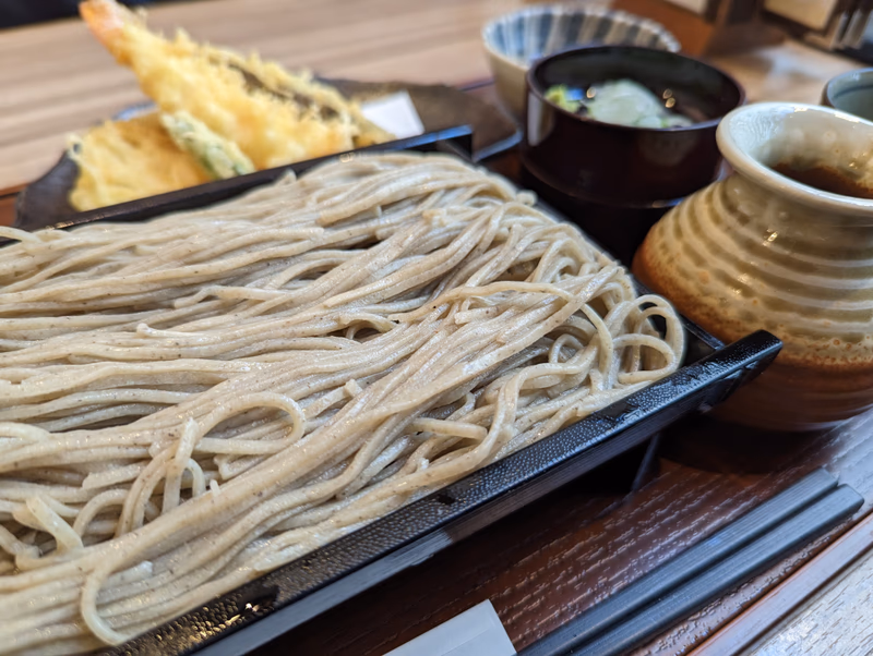 A photo of a bowl of soba noodles with tempura shrimp, a small bowl of miso soup, and a pitcher of miso soup.