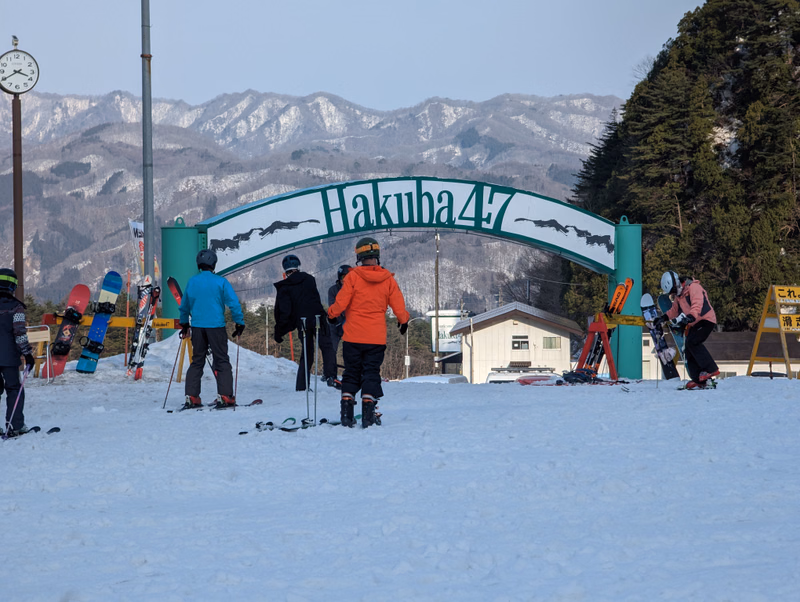 Several skiers are visible on a snowy slope in front of a large archway sign that reads "Hakuba 47" with mountains in the background.