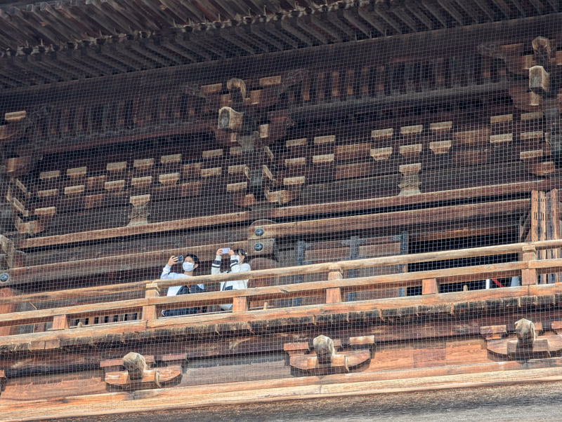 Several people are visible on a wooden balcony of an old temple structure, viewed through a protective mesh.