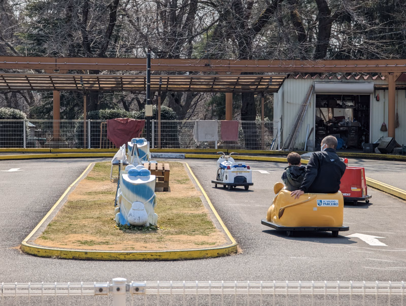 A collection of whimsical sculptures sits on a grassy median in front of a building, with a yellow vehicle and people nearby.