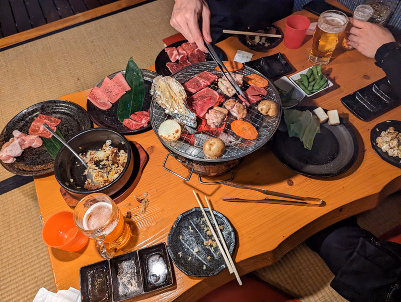 A group is gathered around a wooden table featuring a hot pot grill filled with various cuts of meat and vegetables, with beer glasses nearby.