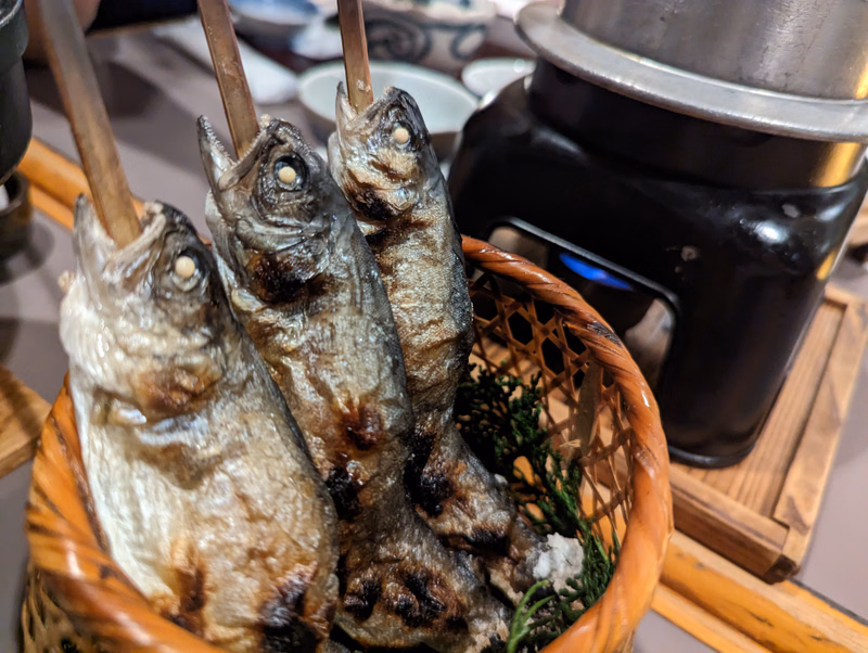 Several dried fish are displayed in a woven basket next to a black grill.