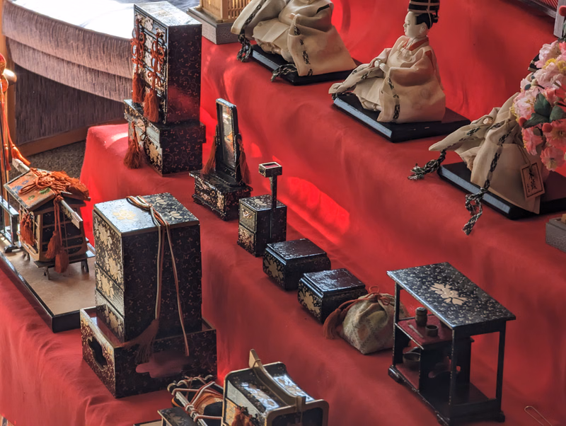 A collection of traditional Japanese dolls and decorative boxes are displayed on a red cloth-covered table.