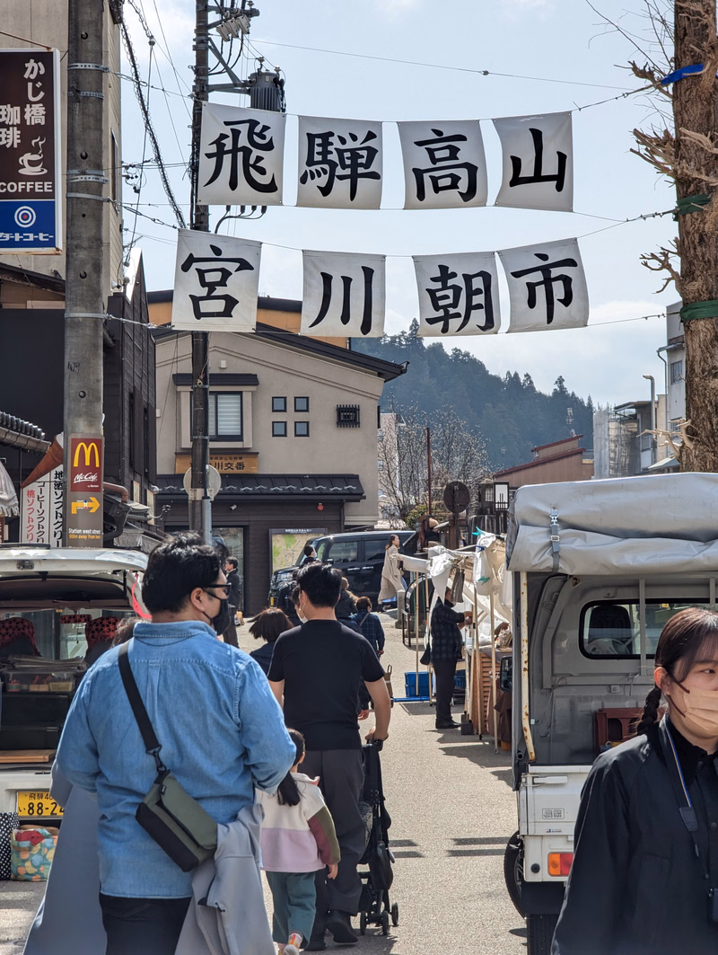 A street scene in a Japanese town featuring large white banners that read "飛騨高山" and "宮川朝市".