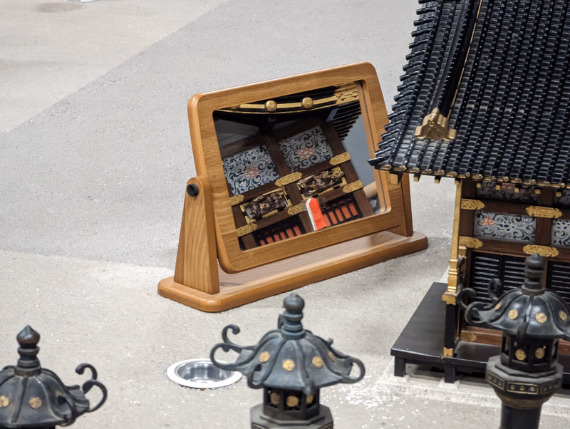 A wooden display box containing a miniature house sits next to a larger, detailed miniature Japanese-style building with a black tiled roof.