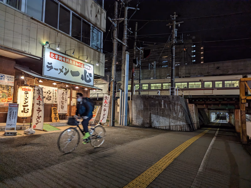 A nighttime scene in an urban area with a cyclist, a building with Japanese signage, and a train passing through a tunnel.