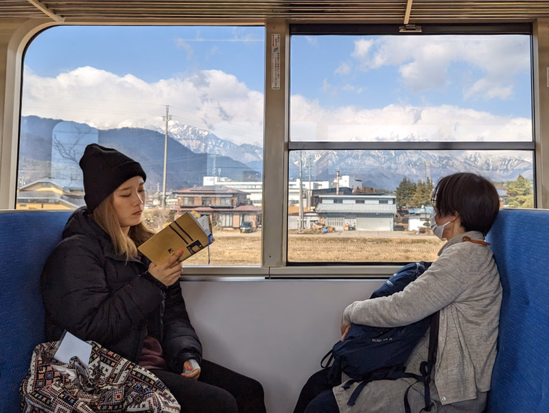 Two people sit inside a train looking out the window at a view of mountains and buildings under a cloudy sky.