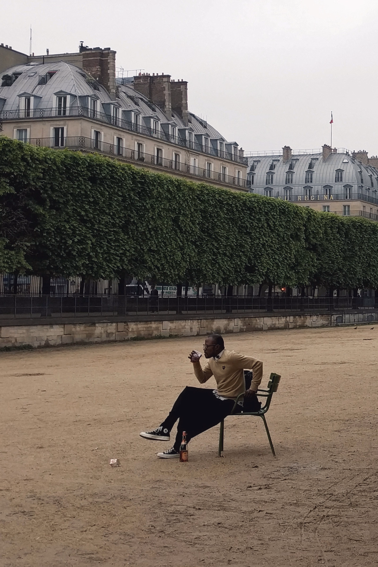 A man in a light brown jacket sits on a metal chair on the sand, with large buildings covered in ivy in the background.